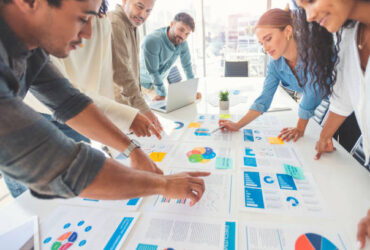 Multi racial group of people working with Paperwork on a board room table at a business presentation or seminar. The documents have financial or marketing figures, graphs and charts on them. People are pointing to different documents. There is a laptop on the table